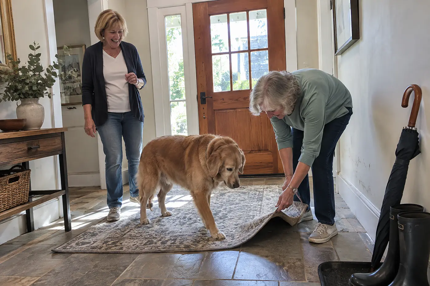 An older woman smooths the edge of a nonslip rug beneath her dog's paws in an entryway.