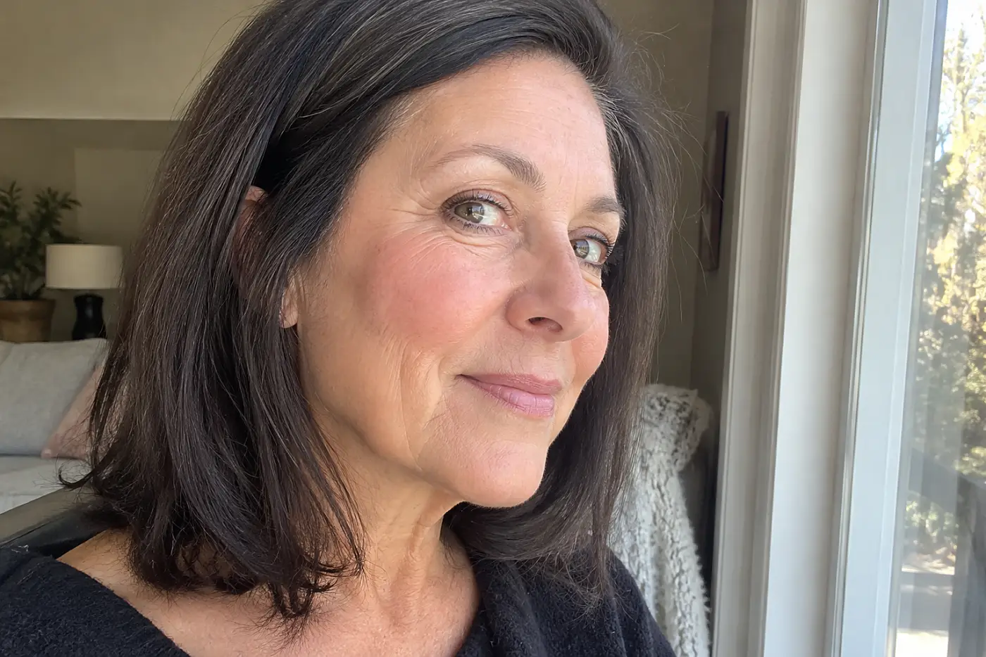Woman turning her cheek toward daylight beside a living-room window after applying blush.