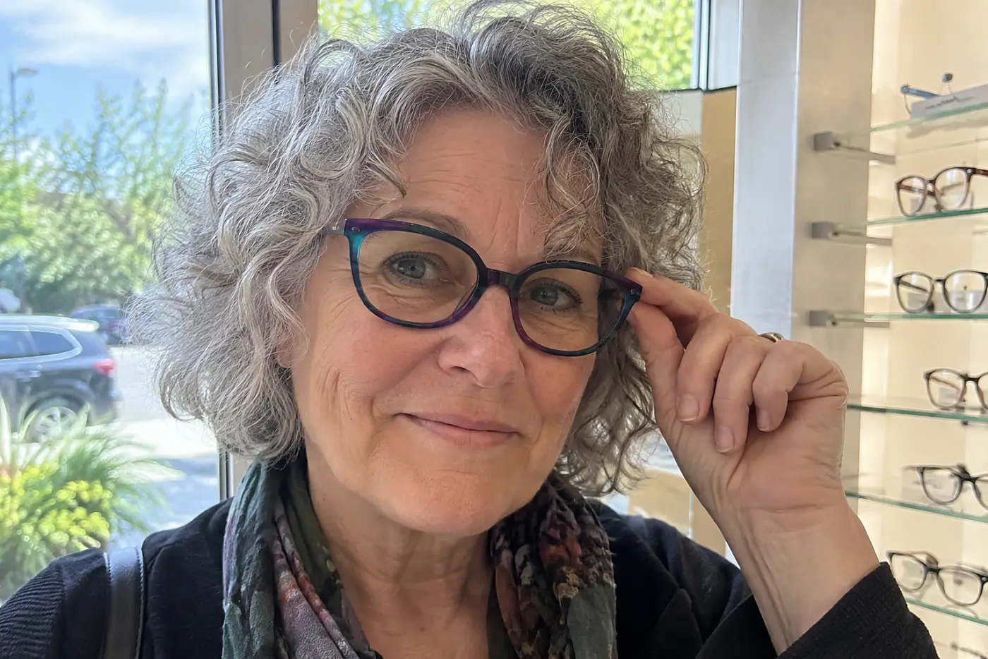 Woman trying on jewel-toned glasses beneath a shop window’s daylight.