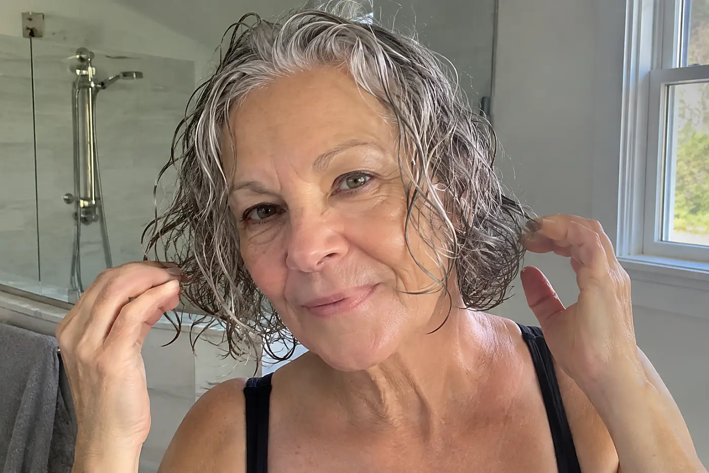A woman works curl cream through her wet bob beside a bathroom sink.