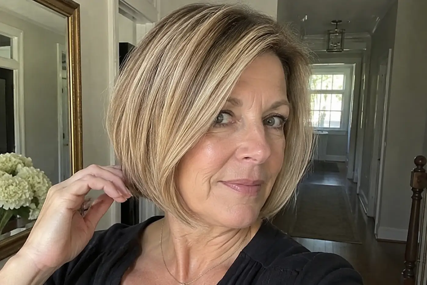 A woman sweeps the front of her cropped bob across her cheek near a hallway mirror.