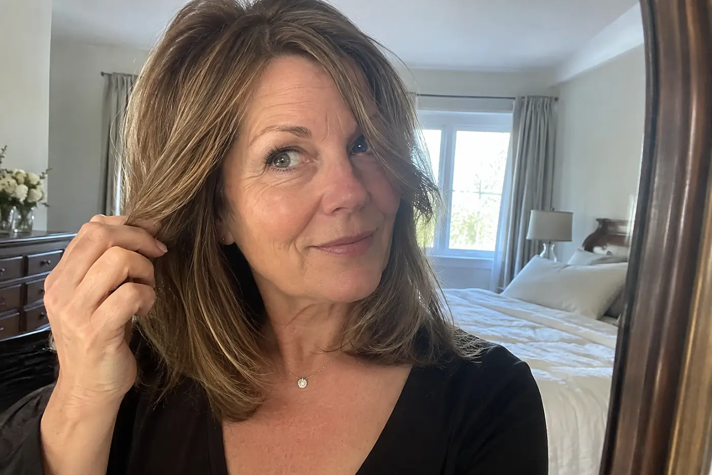 A woman pulls a face-framing piece beside her cheek in a bedroom mirror.
