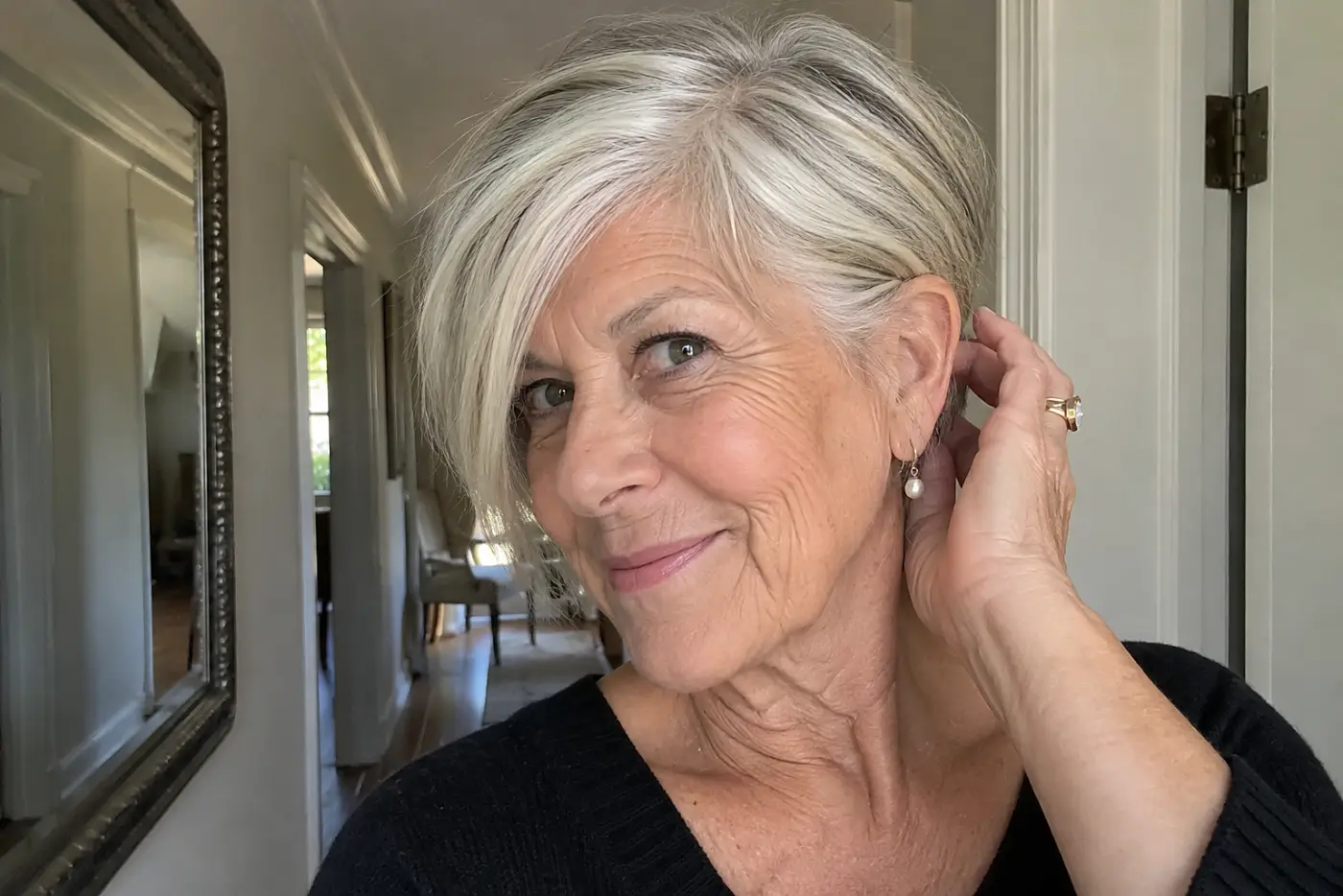 A woman tucks the side of her pixie-bob behind her ear beside a hallway mirror.