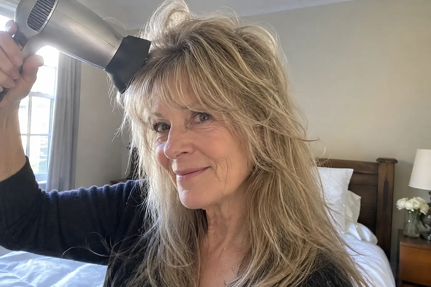 A woman blow-dries the front layers of her long shag in her bedroom.