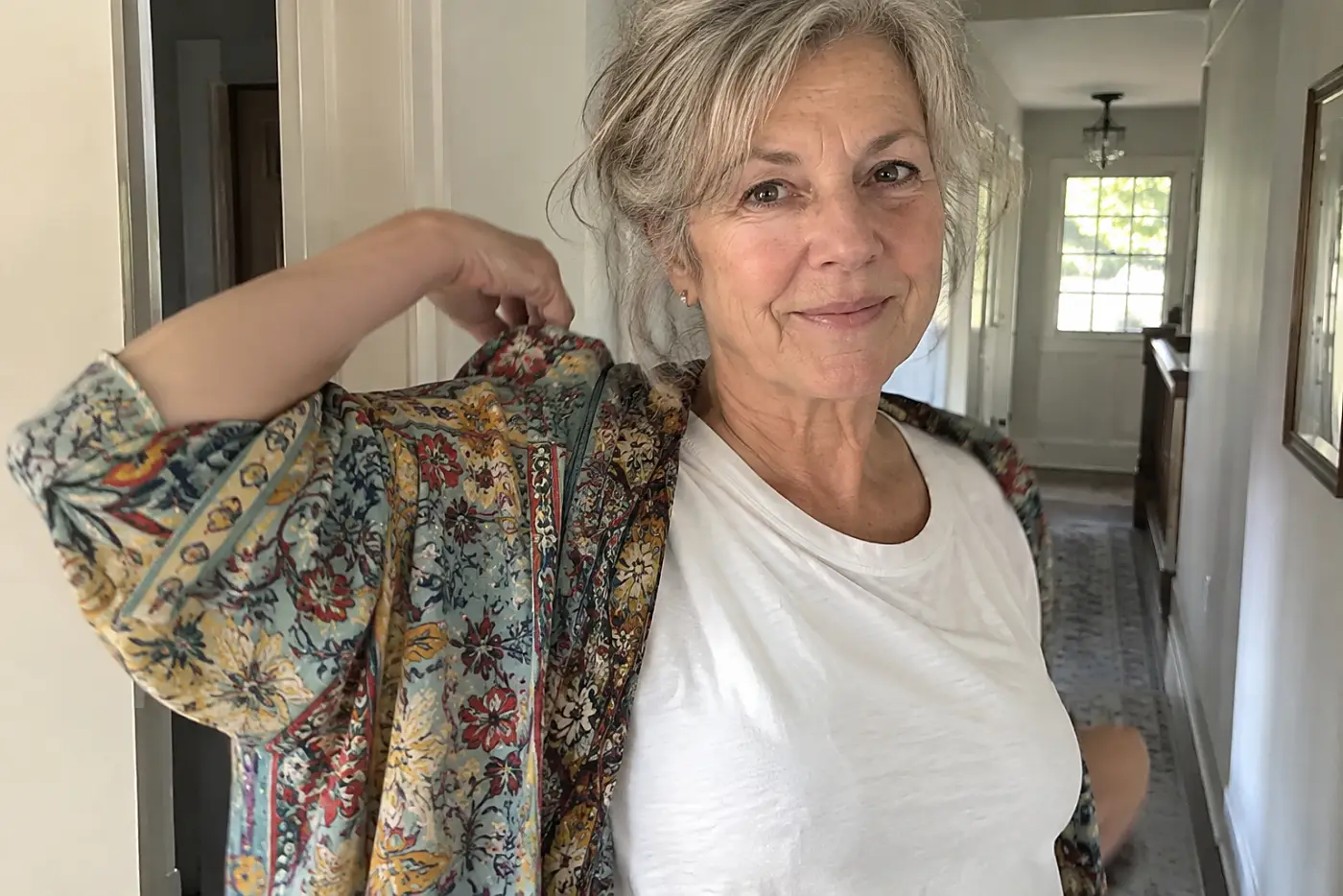 Older woman putting a printed kimono over a plain tee in her hallway.