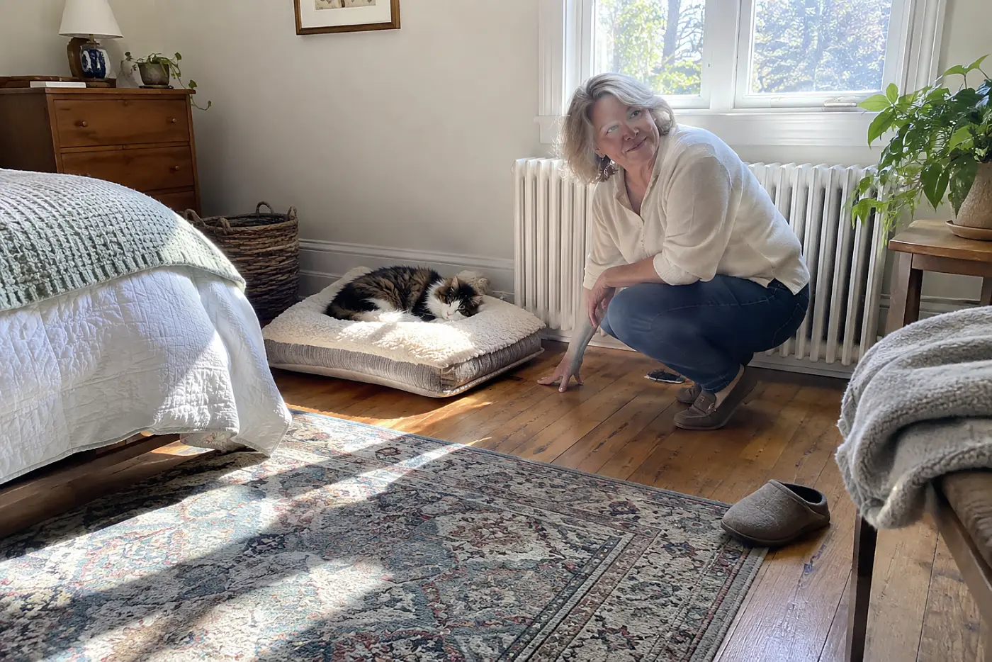 Older woman checking the floor beside a radiator while her cat rests in a guest room.