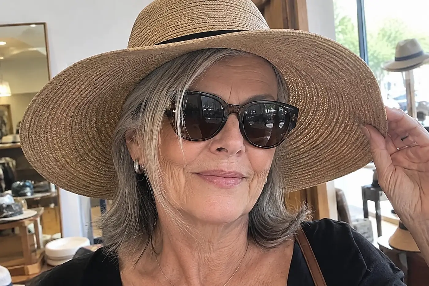 Older woman trying on a wide-brim hat with sunglasses in a shop mirror.