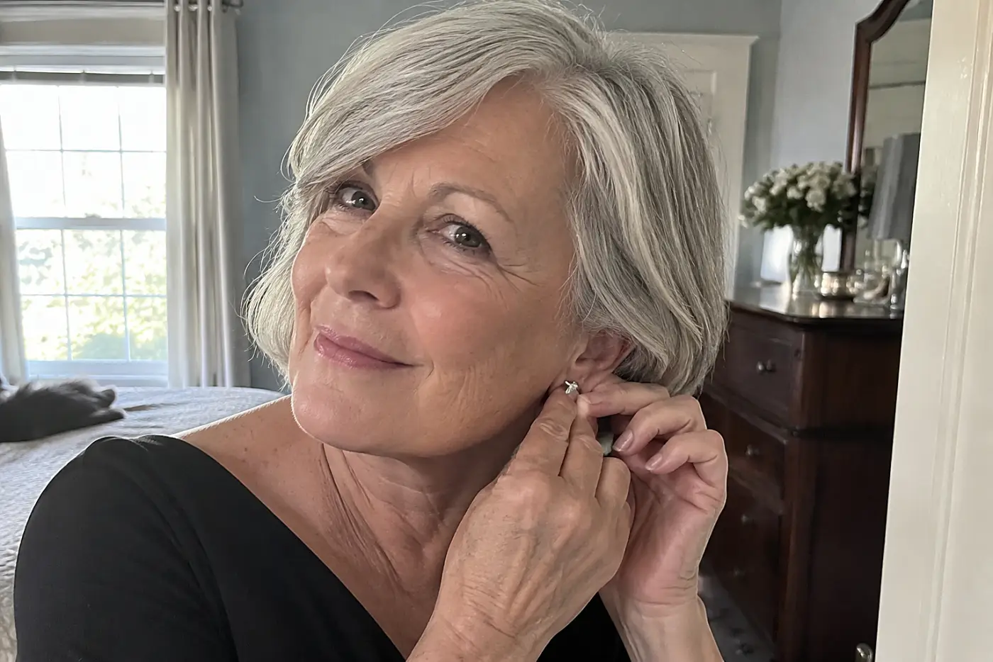 Older woman trying on earrings with a black dress in her bedroom mirror.