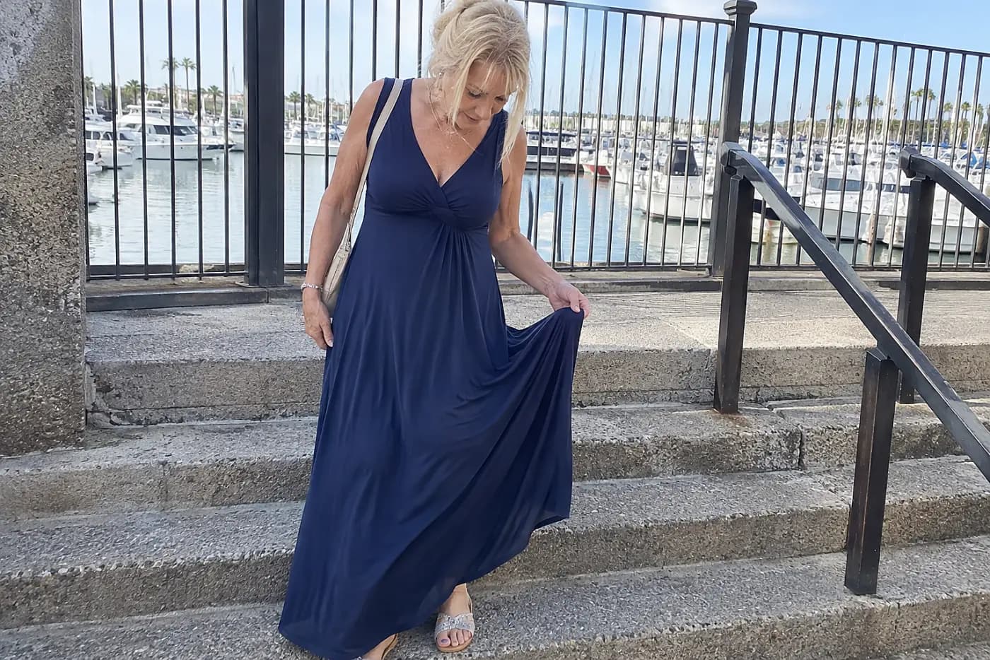Older woman checking the hem of a maxi dress on outdoor steps near a harbor.