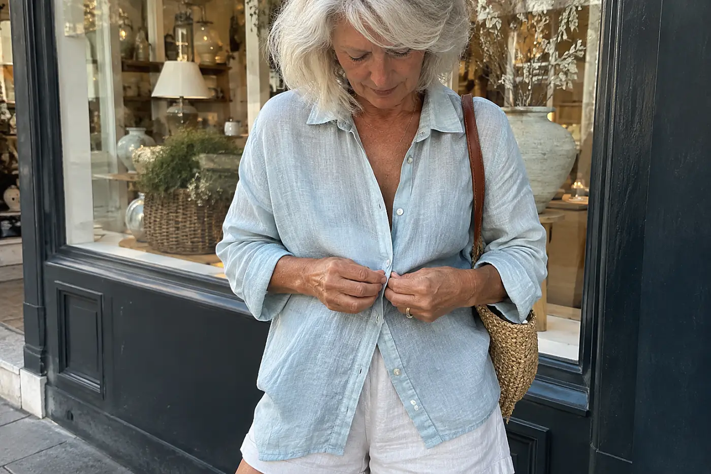 Older woman buttoning a linen shirt over shorts outside a port town shop.