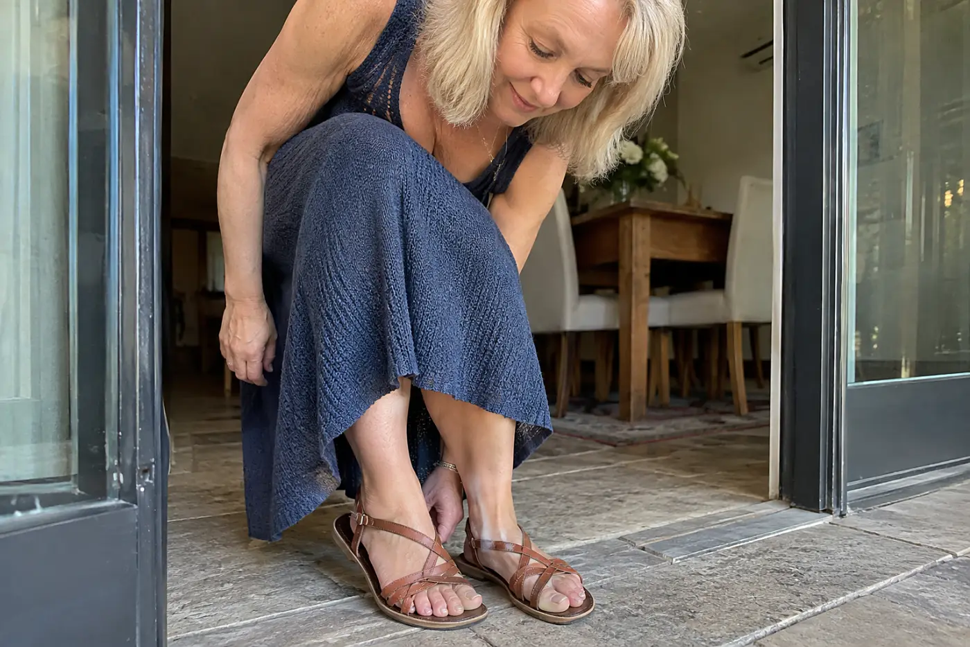 Older woman slipping on sandals with a knit midi dress outside a dining room.