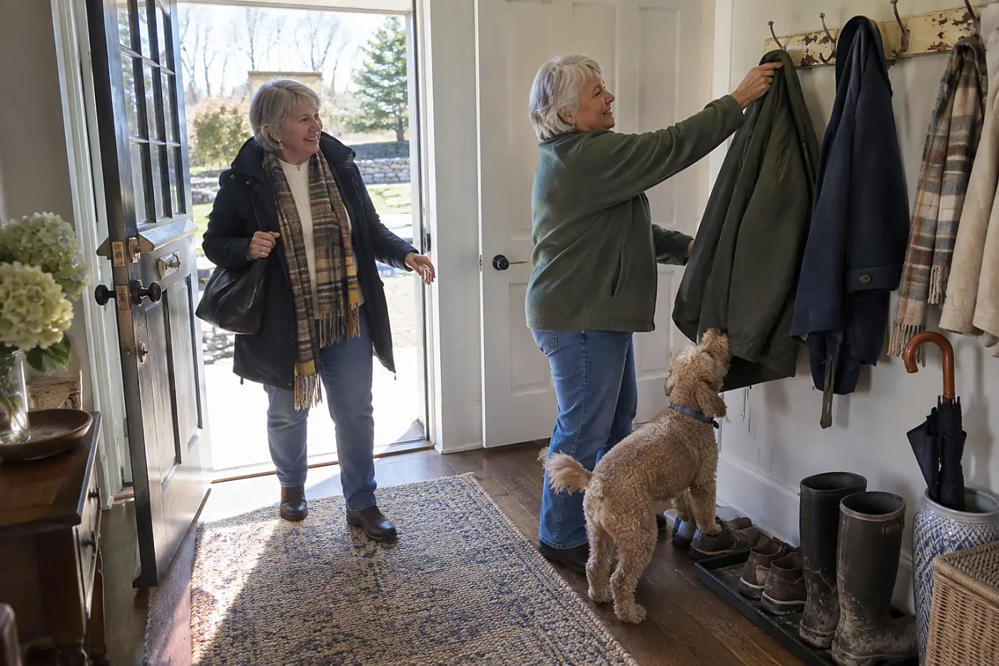 Older woman hanging her coat in an entryway while her dog sniffs its pocket.