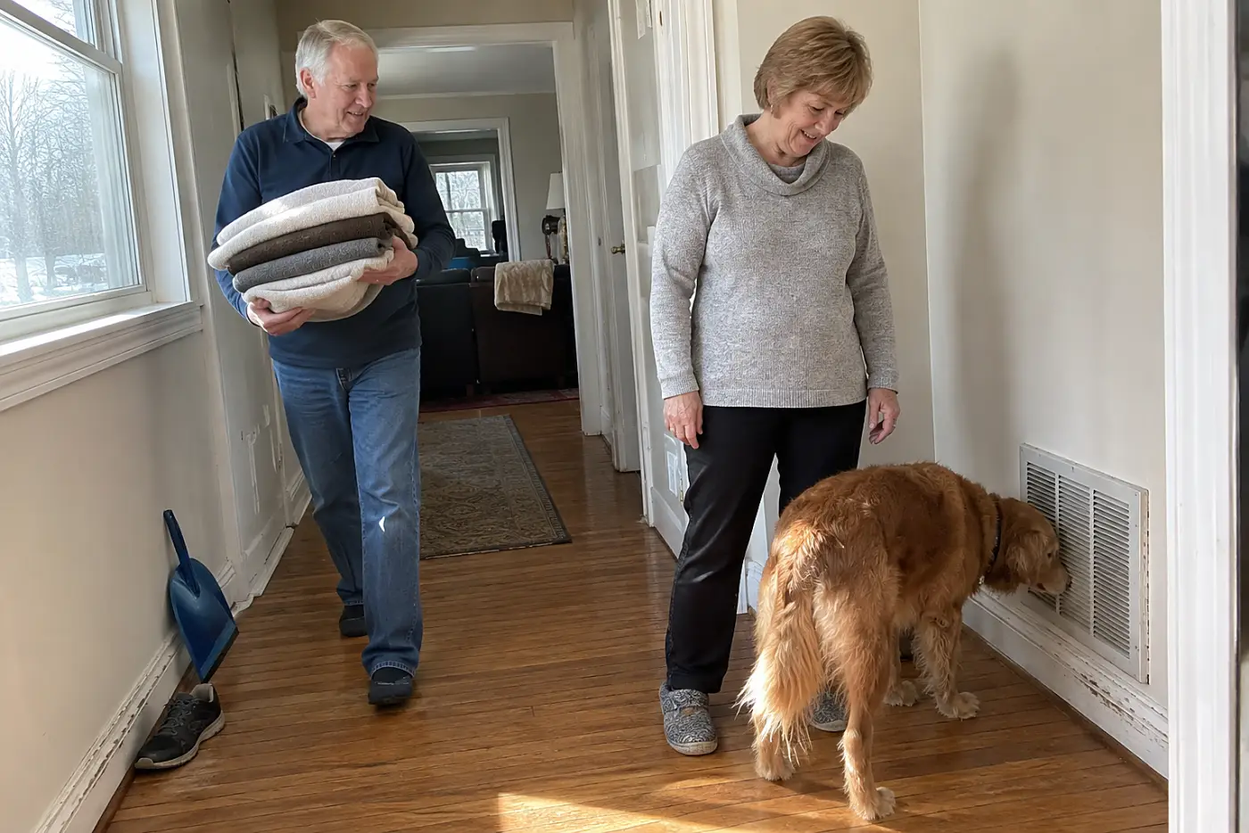 Older woman standing beside her dog in a hallway near a furnace vent.
