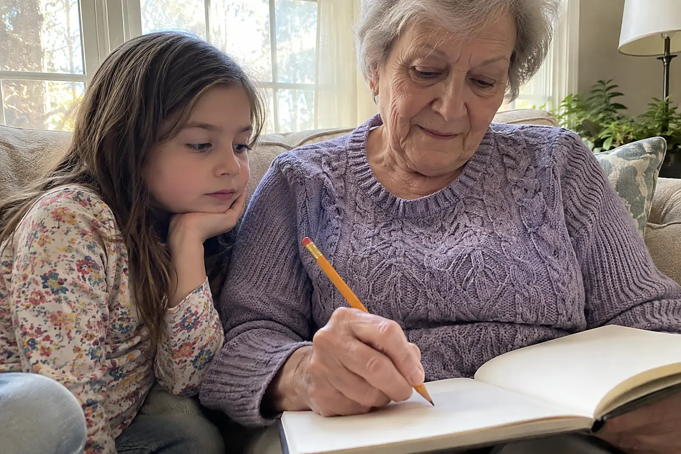 A grandmother writes in a guided journal while her grandchild sits beside her on a sofa.