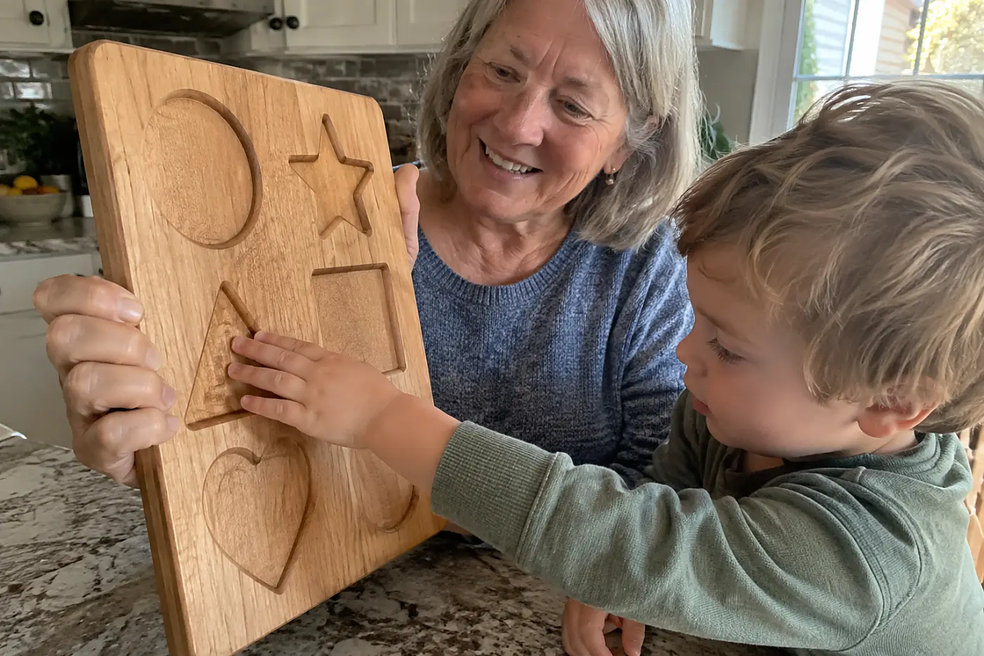 An older woman holds a wooden recipe board beside her grandchild at a kitchen counter.