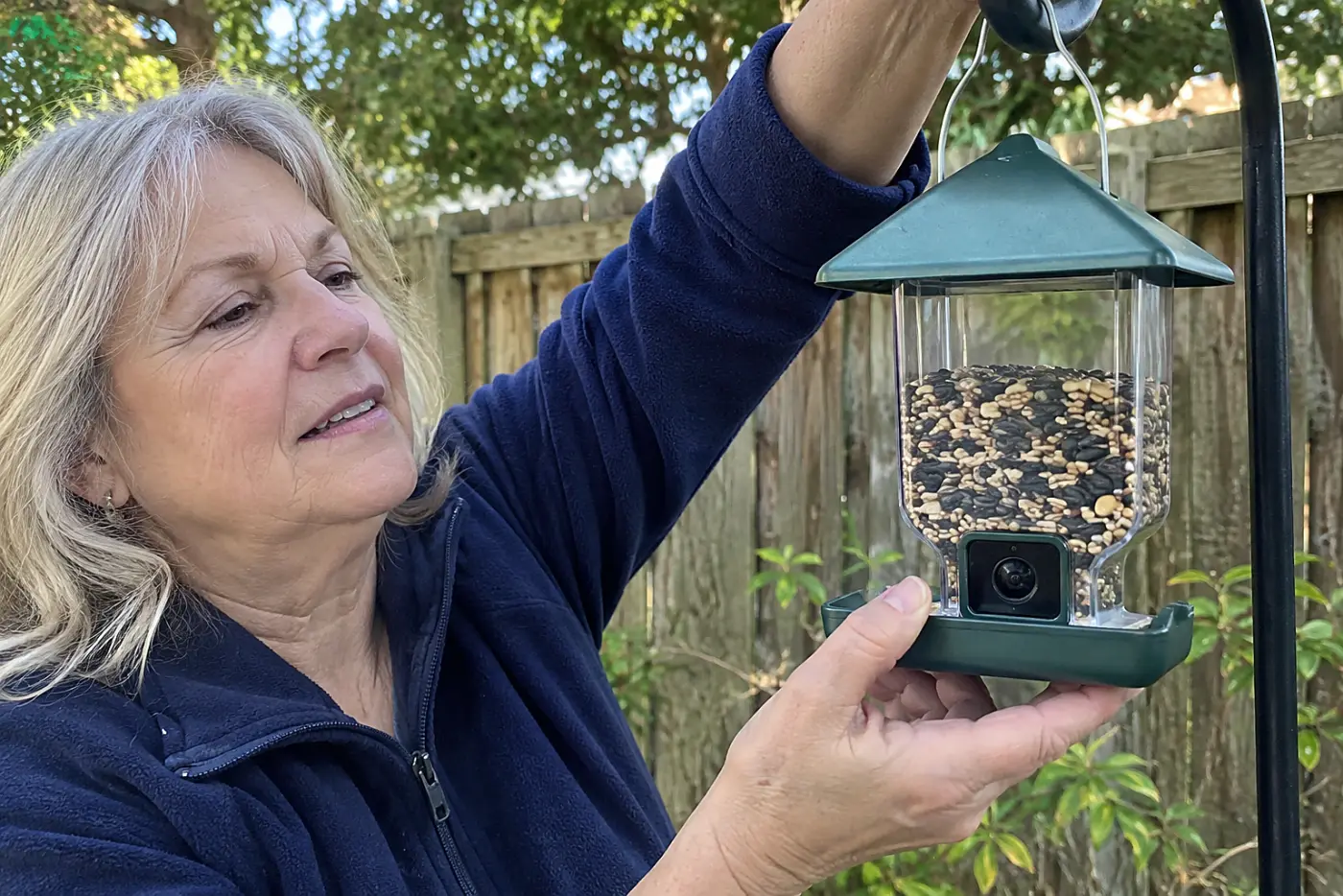 Woman in her sixties hanging a camera bird feeder from a backyard hook.