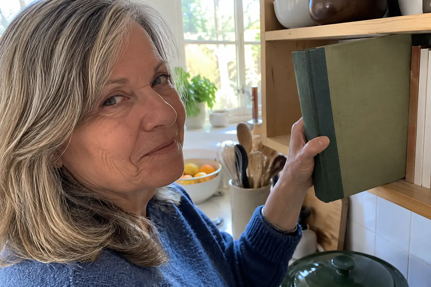 Woman in her sixties taking a cookbook from a shelf beside her kitchen counter.
