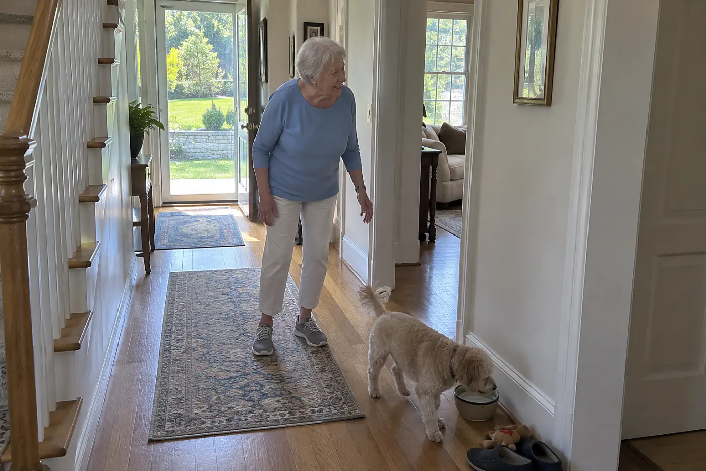 Older woman watching her dog cross a hallway toward a water bowl.