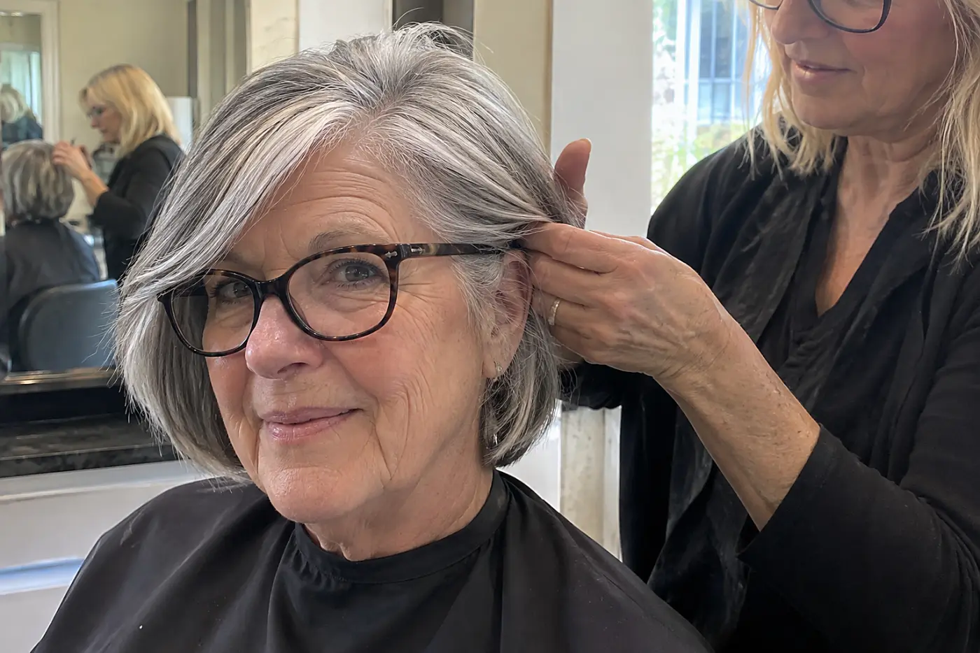 Stylist checking the hair behind an older woman’s glasses arm in a salon.