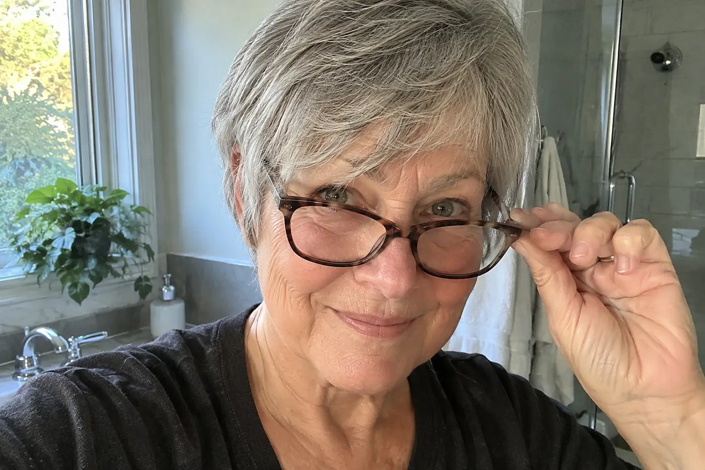 Older woman putting on her glasses beneath short bangs at a bathroom sink.