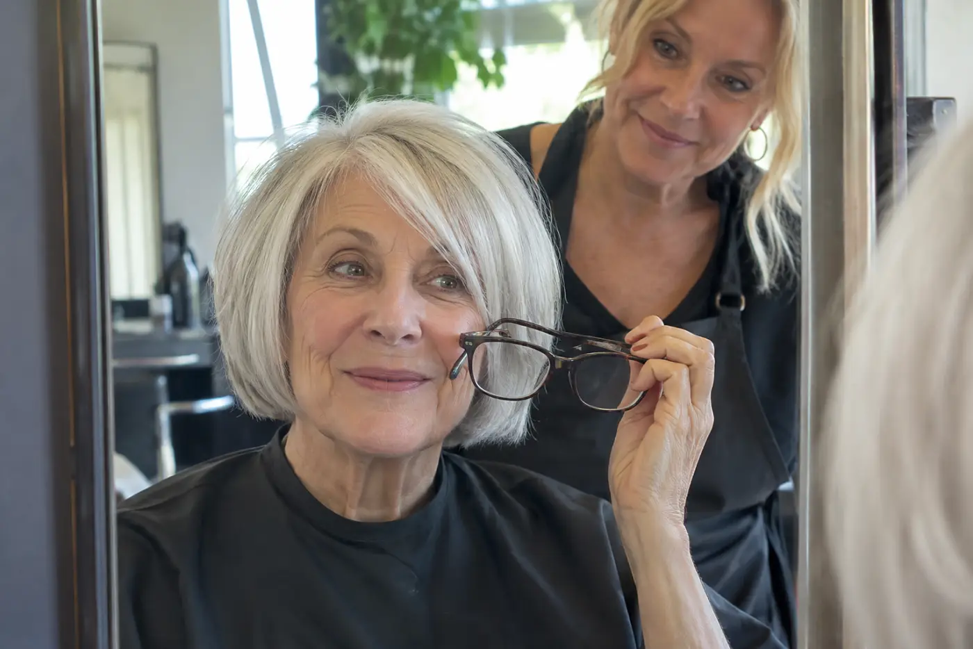 Older woman holding her glasses beside her face in a salon mirror while a stylist watches.