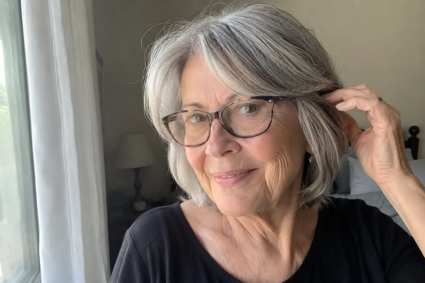 Older woman moving curtain bangs away from her frames beside a bedroom window.