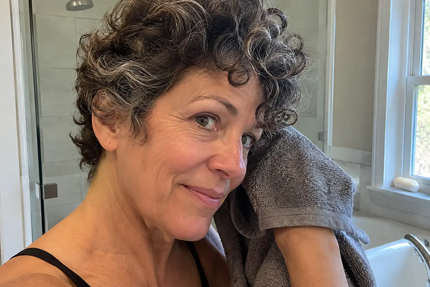 Curly-haired woman drying her pixie with a microfiber towel in a bathroom.