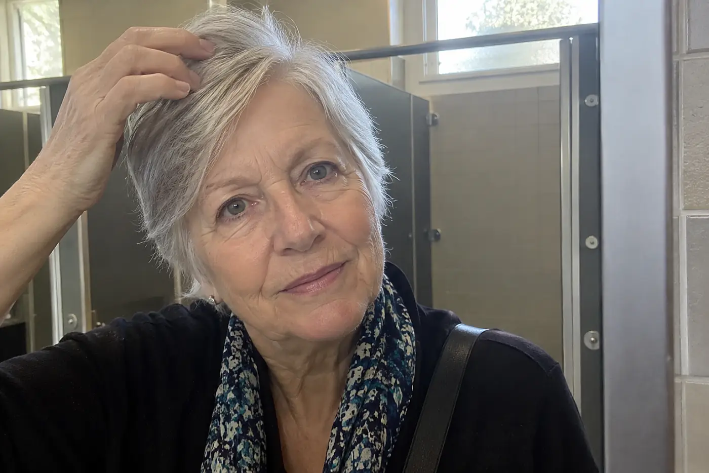 Older woman changing her hair part in a train-station restroom mirror.