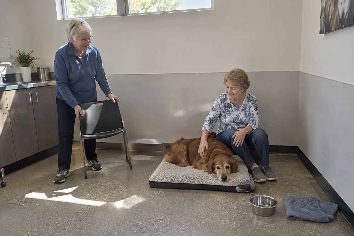 An older woman sits beside a pet blanket in a veterinary exam room, her hand resting on its edge.