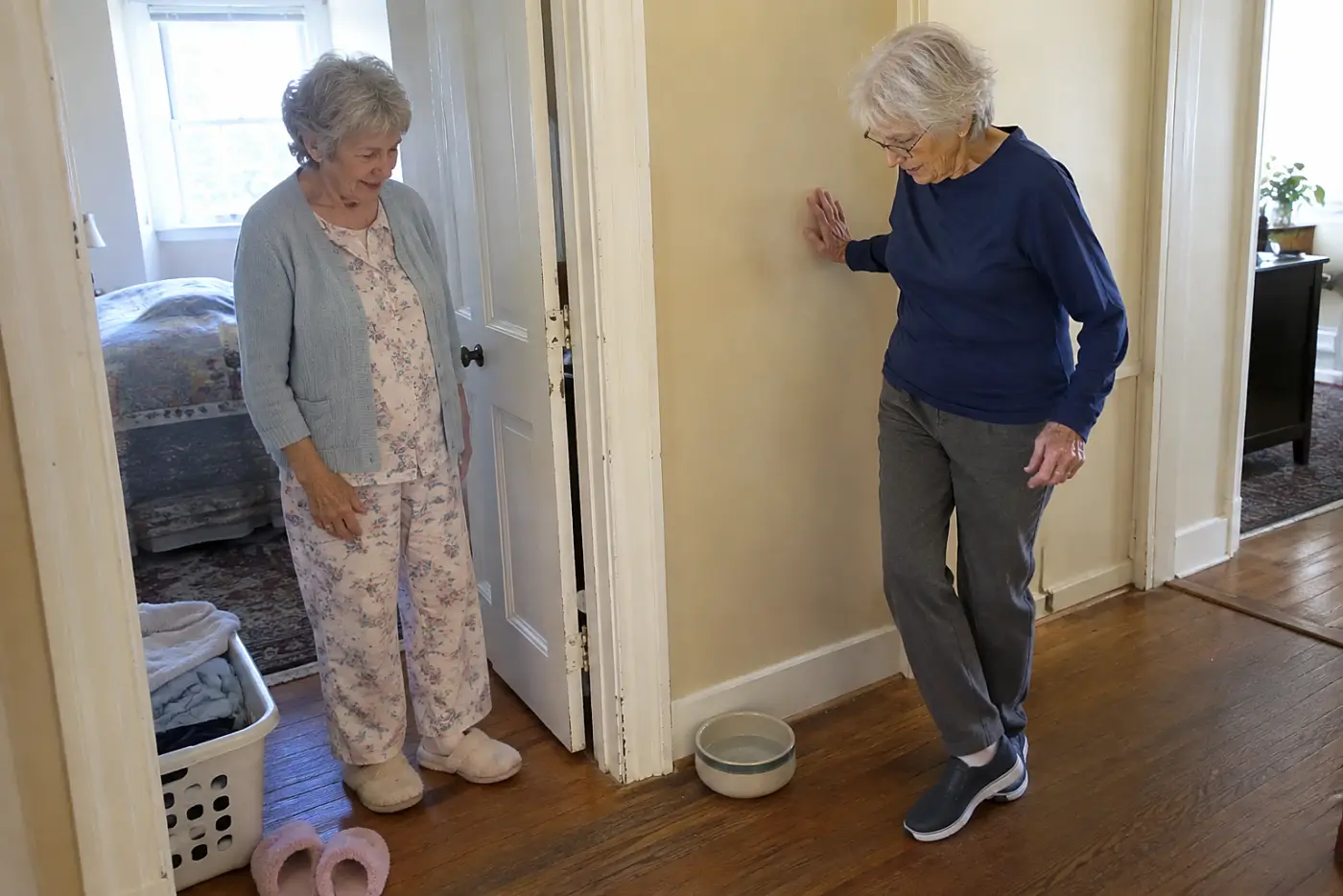 An older woman opens her bedroom door and looks toward a water bowl in the hall.