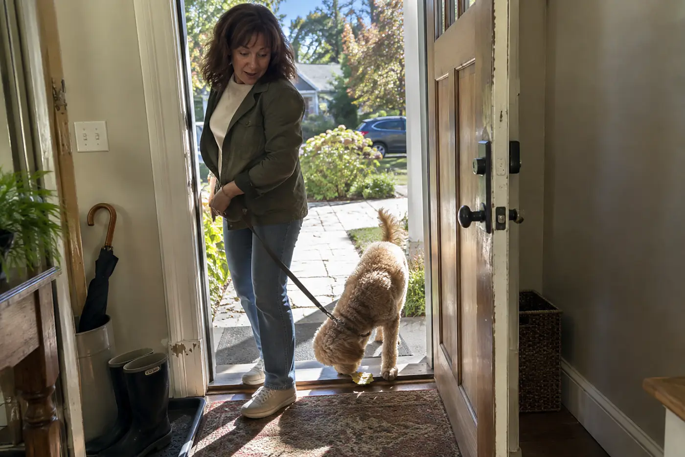 Woman in her fifties holding her dog’s leash while he steps across the front-door mat.
