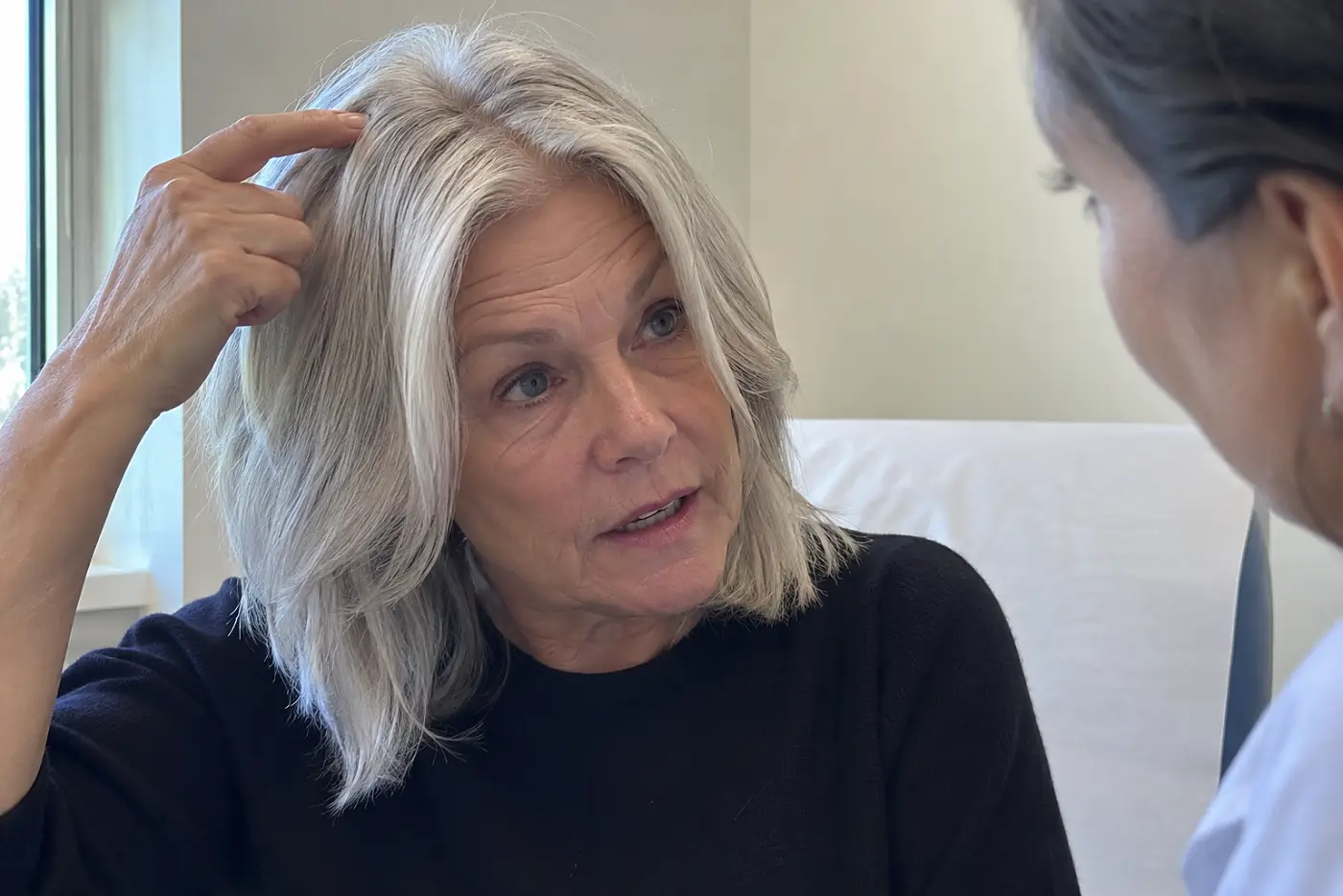 An older woman points to her part while speaking to a dermatologist in an exam room.