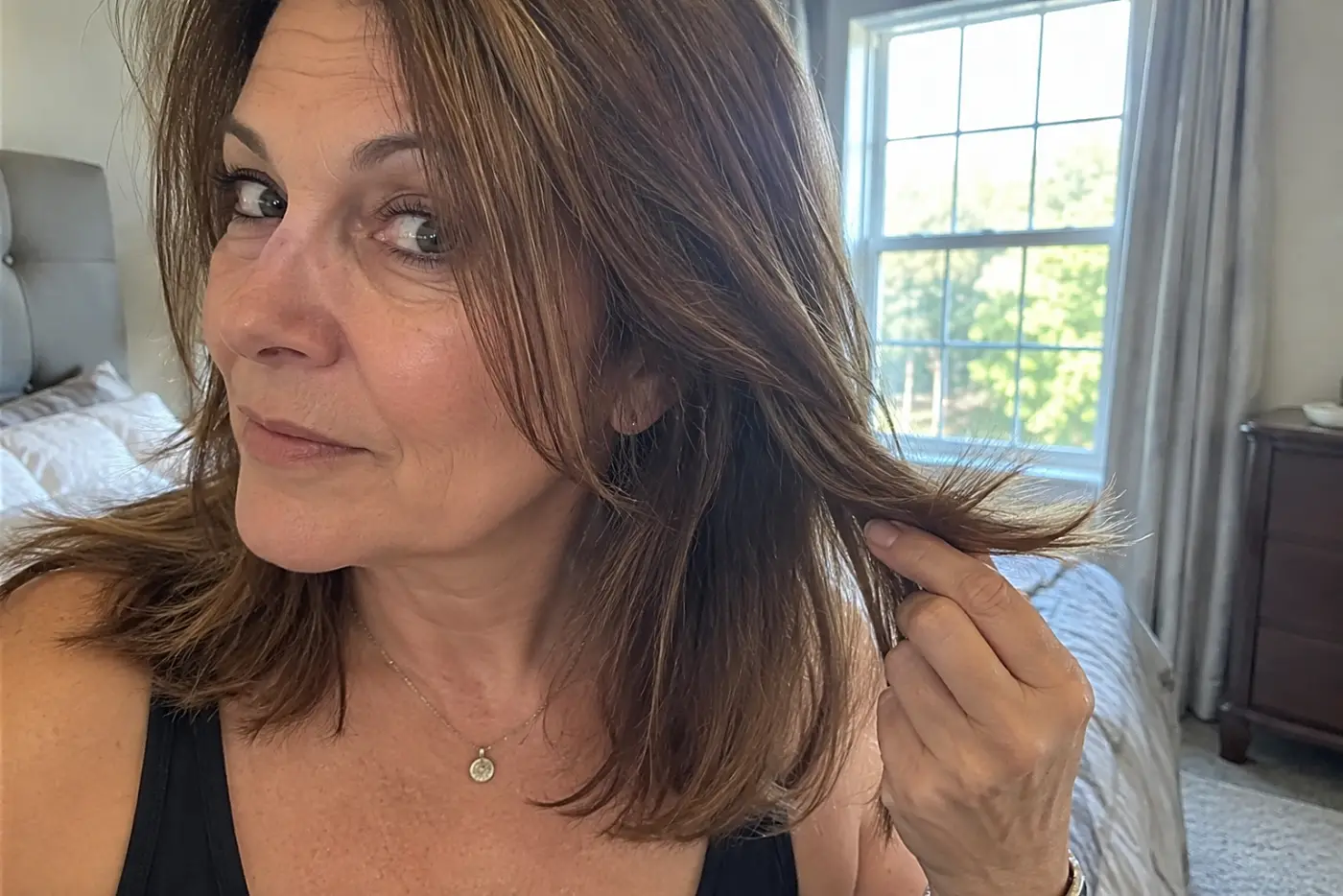 An older woman examines the ends of her hair after brushing it in a bedroom.
