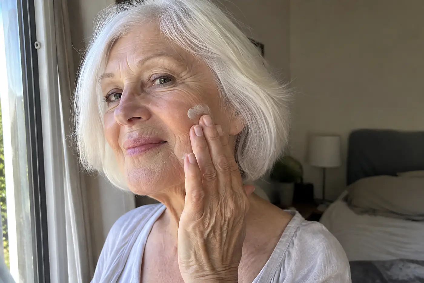 Older woman applying moisturizer beside an open bedroom window.