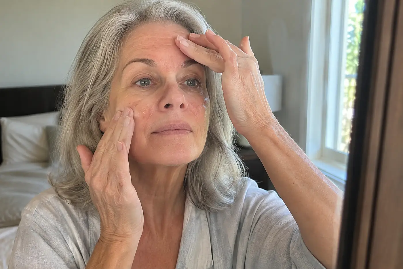 Older woman applying serum in a bedroom mirror.