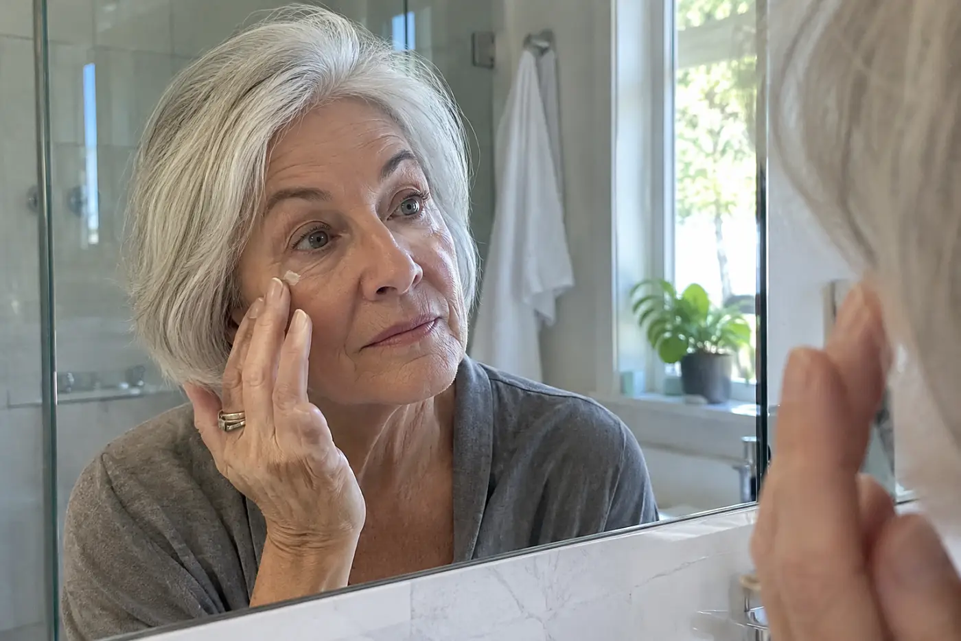 Older woman dabbing eye cream beside her bathroom mirror.