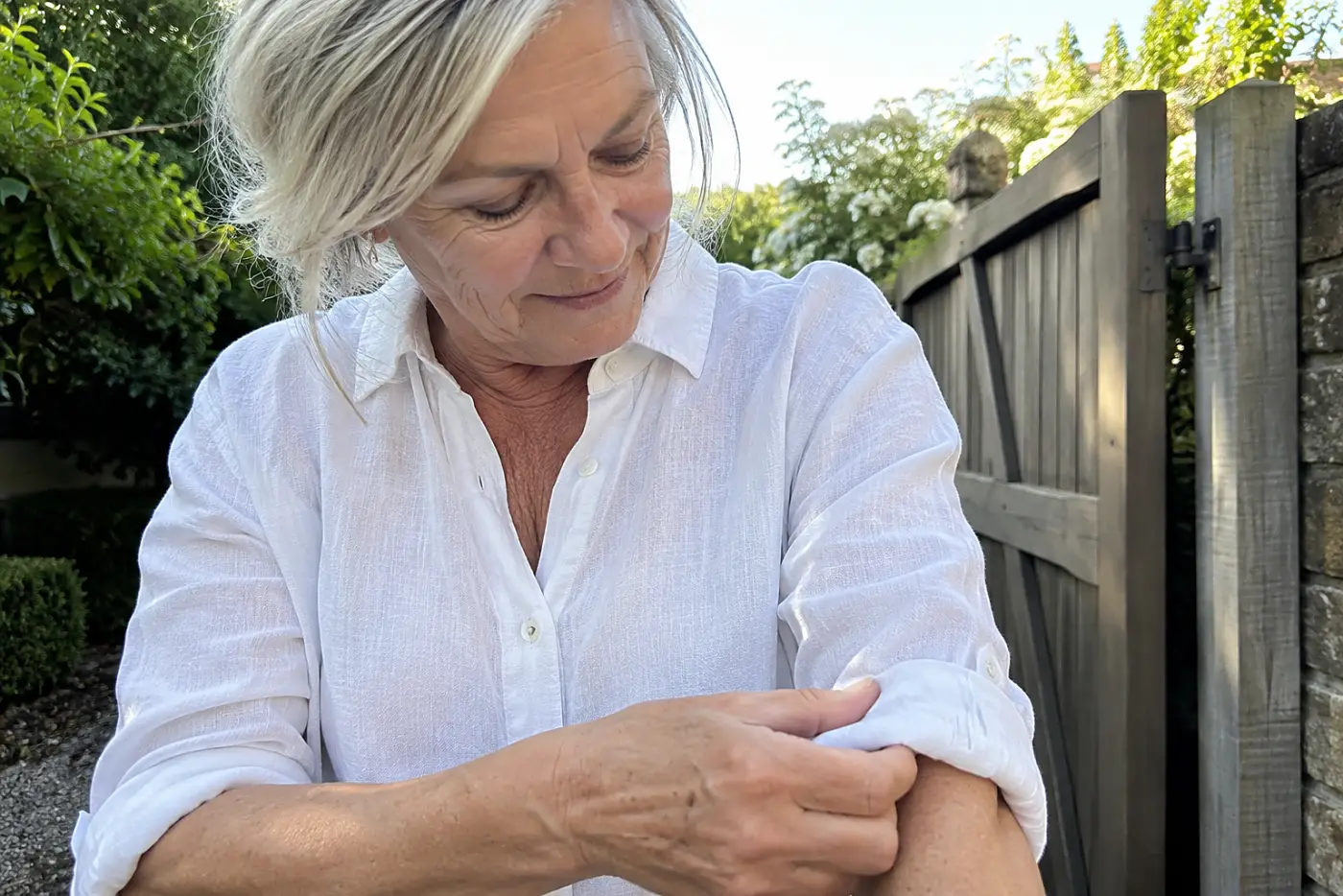 A woman rolls the sleeves of her linen shirt beside a garden gate.