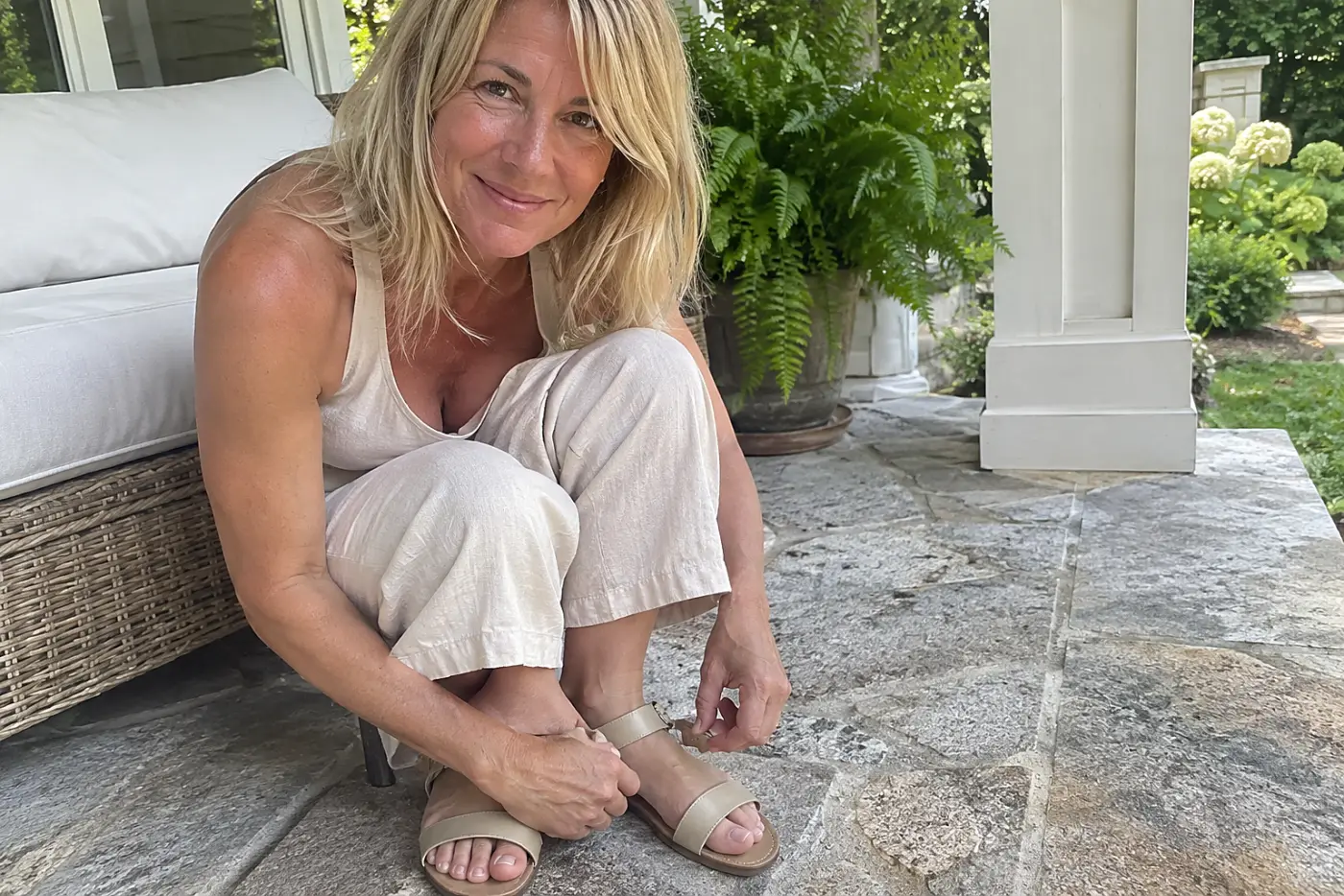 A woman fastens flat sandals beneath matching linen separates on a porch.