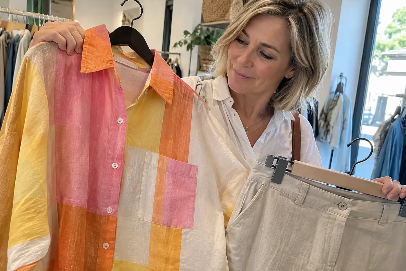 A woman holds a colorful linen shirt beside neutral trousers in a boutique.