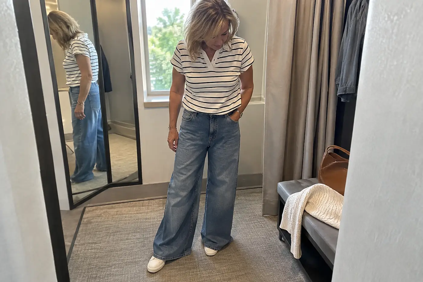 A woman checks the hem of wide-leg jeans with a striped polo in a fitting room.