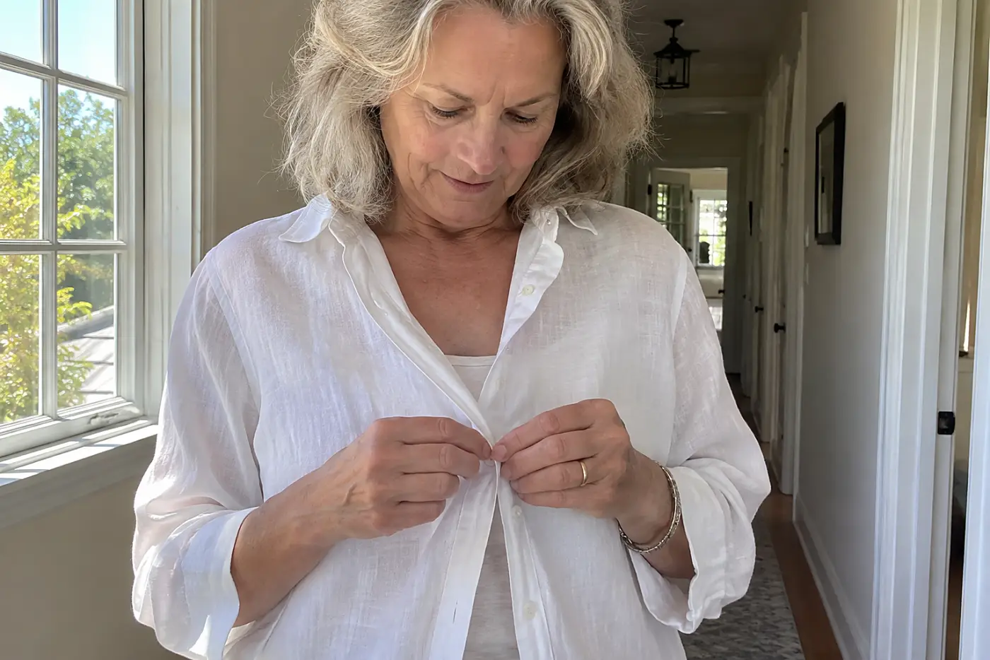 A woman buttons a white linen shirt over a tank in a sunlit hallway.