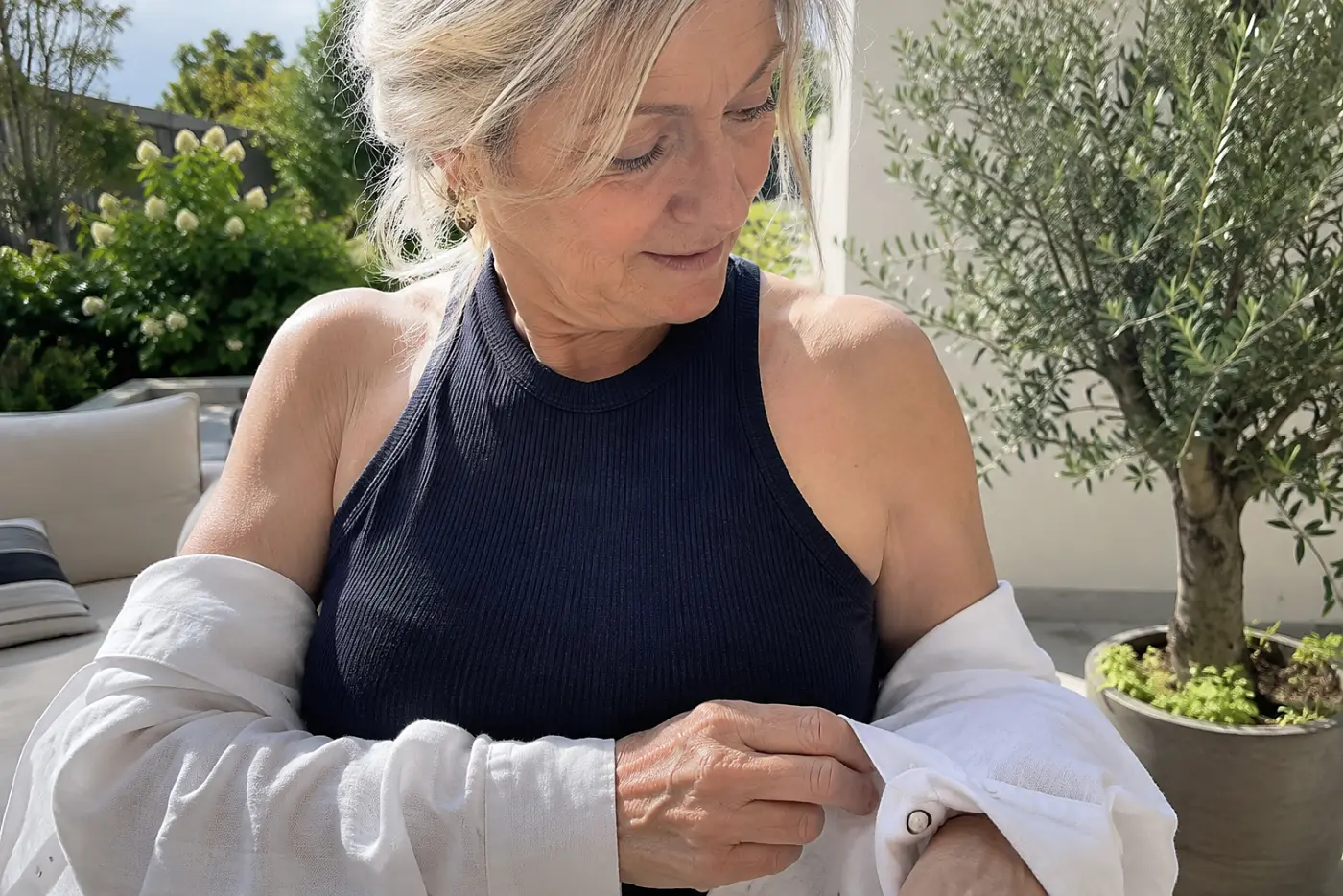 A woman takes off her linen shirt to reveal a tank on a sunny patio.