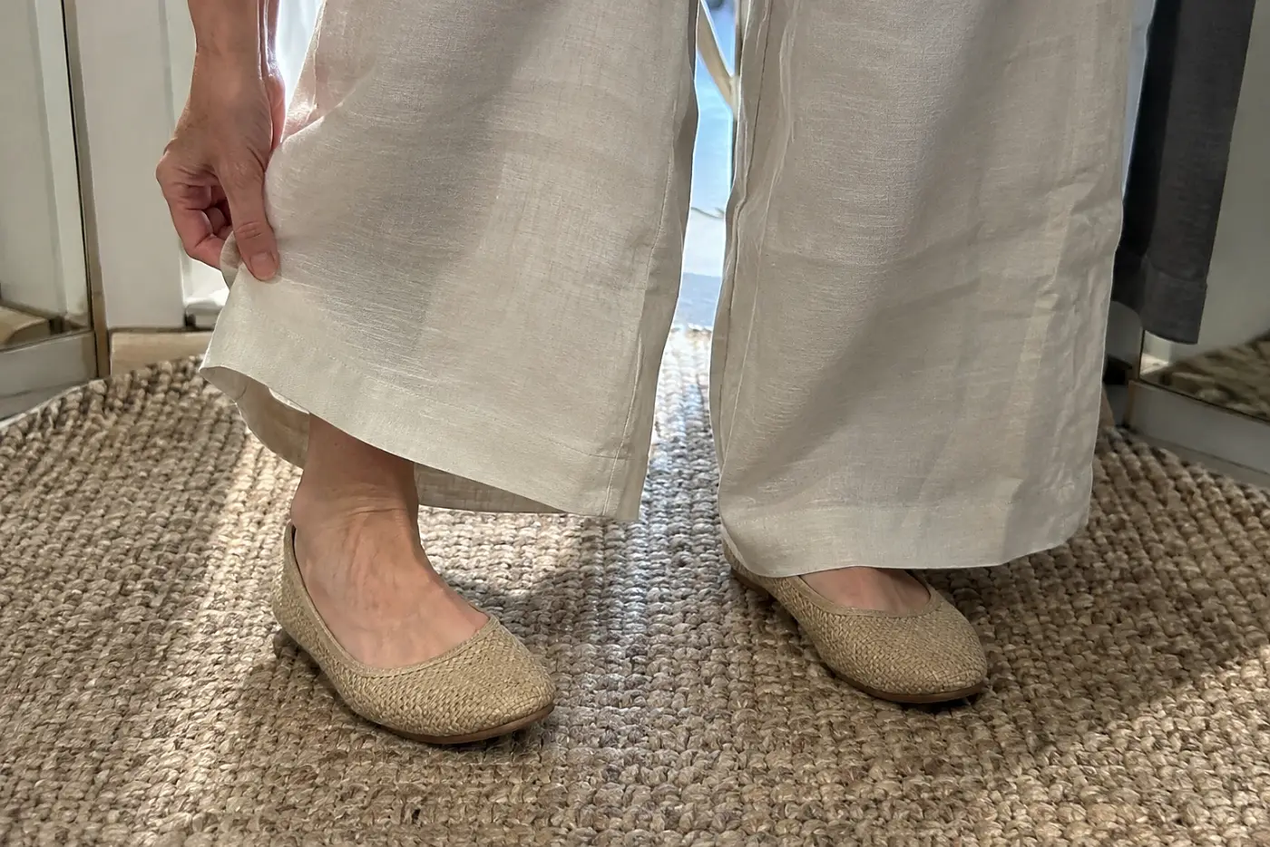 A woman compares the hem of wide-leg linen pants with her shoes in a fitting room.