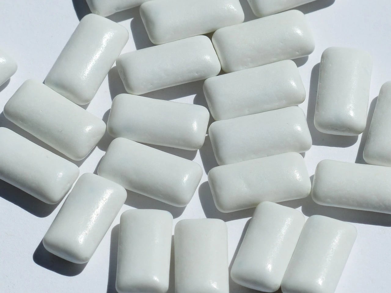 Close-up of many white rectangular candy-coated chewing gum pieces scattered on a white surface in bright light
