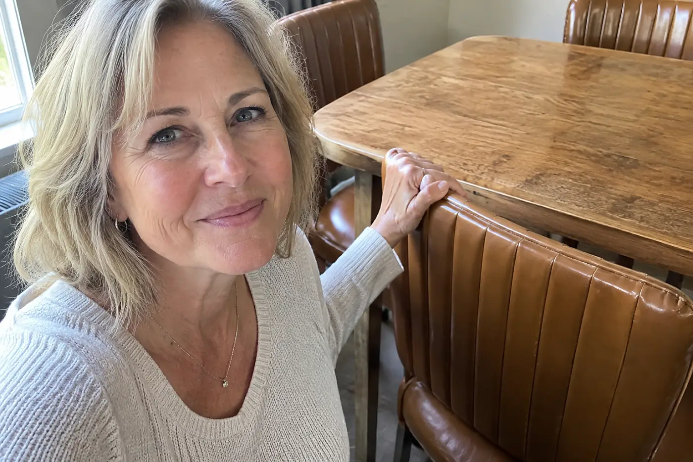 A smiling woman in her sixties touches a brown padded vinyl kitchen chair.