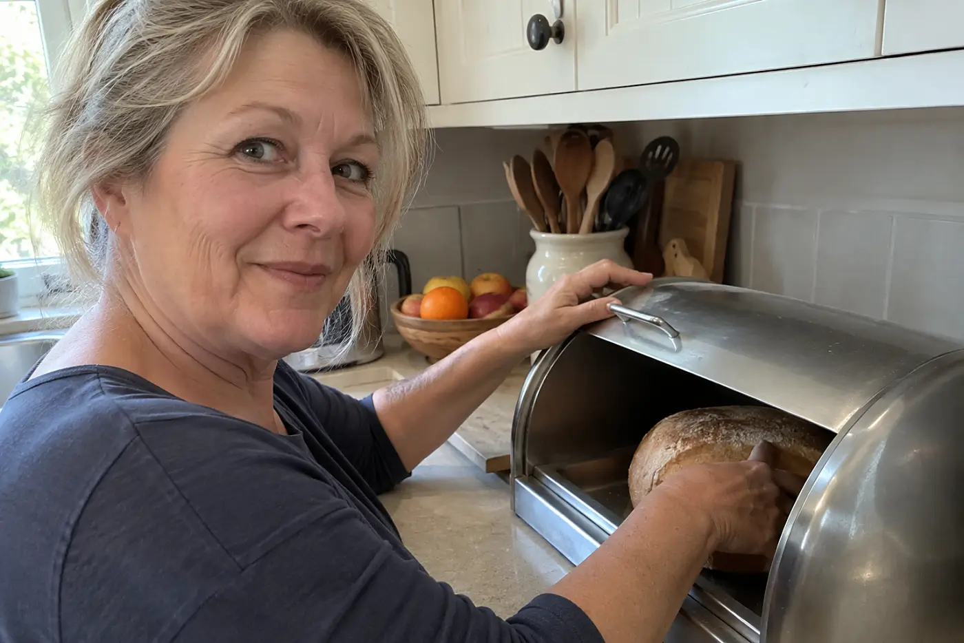 A woman in her sixties reaches for a loaf of bread inside a metal bread box.