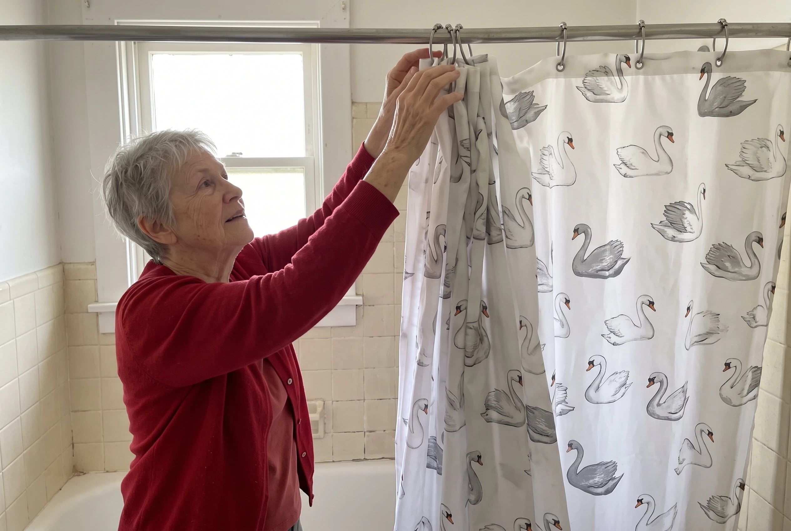 An older woman pulls a swan-print shower curtain across her bathtub.