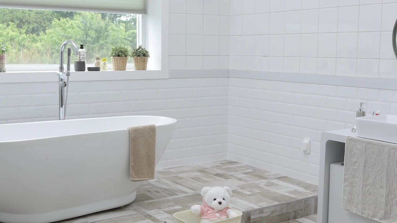 White freestanding bathtub with chrome floor-mounted faucet beside a window in a white-tiled bathroom with a teddy bear