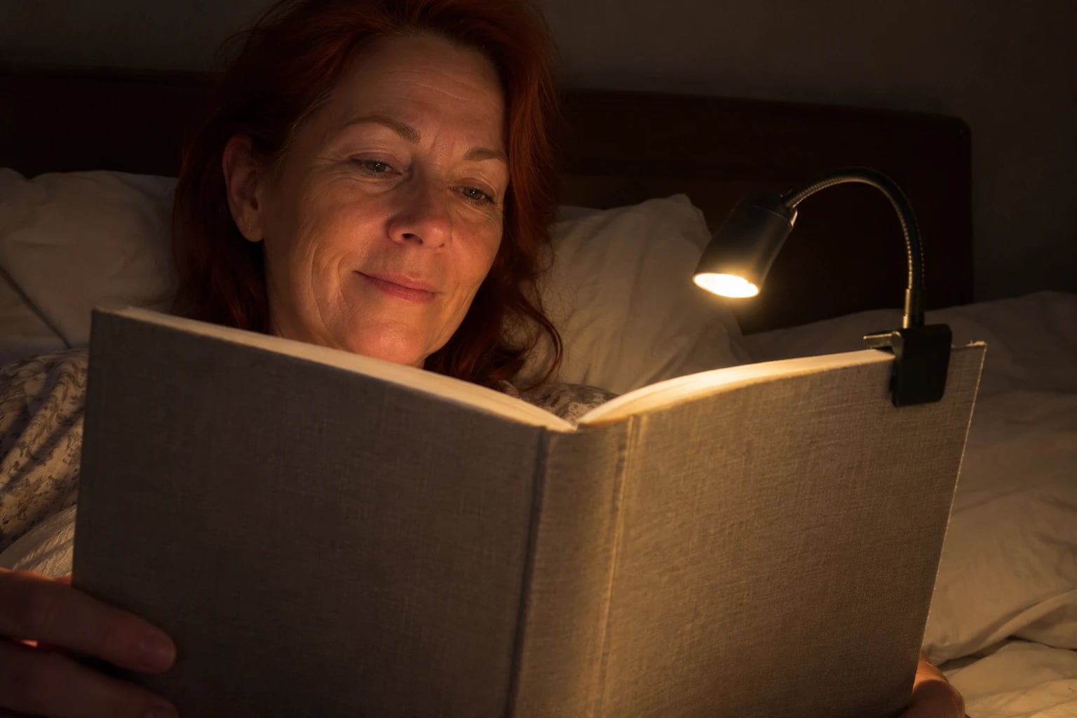 In a dim bedroom at night, a woman in her fifties sits up against the pillows reading a book with a plain cloth cover, a small clip-on gooseneck light gripping its top edge and lighting only the page.
