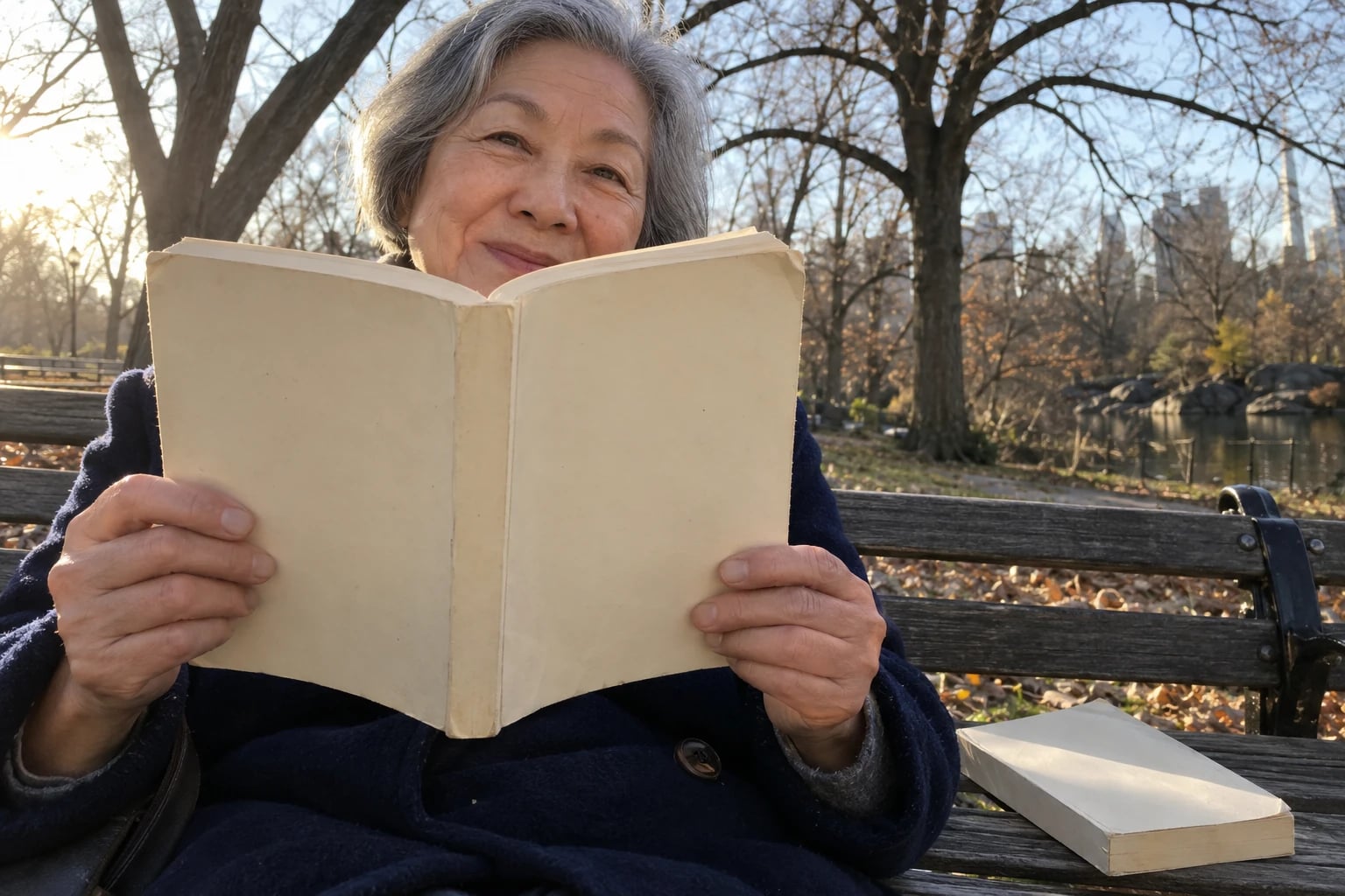 On a park bench under bare autumn trees, a woman in her sixties reads a worn paperback with a plain cream cover and no lettering, a second identical-looking copy resting on the bench beside her.